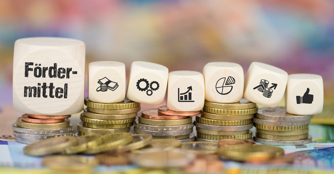 Kleine Stapel von Geldmünzen auf denen Würfel liegen. Auf dem ersten Würfel steht das Geld Fördermittel, auf den restlichen sind Symbole für Finanzen, Wirtschaft und Zusammenarbeit abgebildet.